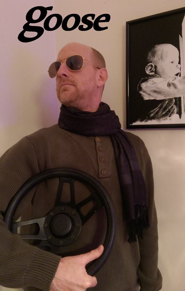 Goose in aviator sunglasses, a scarf, and an olive sweater, cradling a steering wheel under one arm, with the word &ldquo;goose&rdquo; in bold lettering across the top of the photo